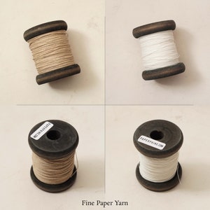 Puede incluir: Dos carretes de hilo de papel fino, uno blanco y otro beige natural, sobre un fondo blanco. Los carretes son de madera y tienen etiquetas con el texto "PAPERPHINE.COM" y "MADE IN THE USA".