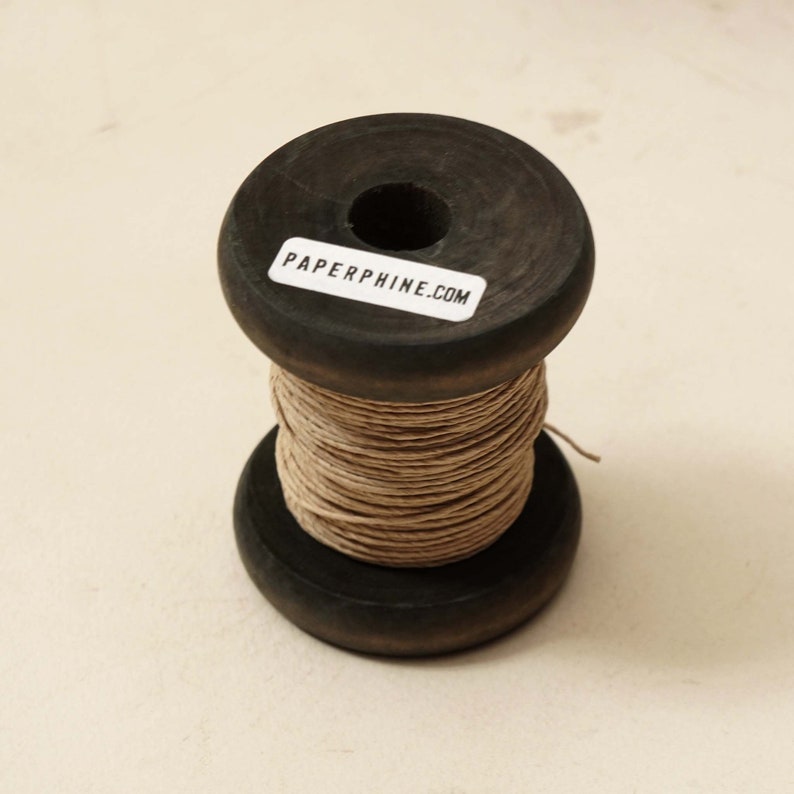 Puede incluir: Una bobina de hilo marr&oacute;n natural enrollada alrededor de una bobina de madera. La bobina tiene una etiqueta que dice "PAPERPHINE.COM".