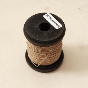 Puede incluir: Una bobina de hilo beige enrollada alrededor de una bobina de madera negra. La bobina tiene una etiqueta que dice "PAPERPHINE.COM".
