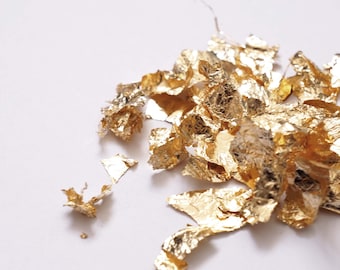 May include: Close-up of scattered gold leaf flakes on a white surface. The flakes vary in size and shape, reflecting light and creating a shimmering effect. Ideal for craft projects, the gold leaf adds a touch of luxury and elegance.