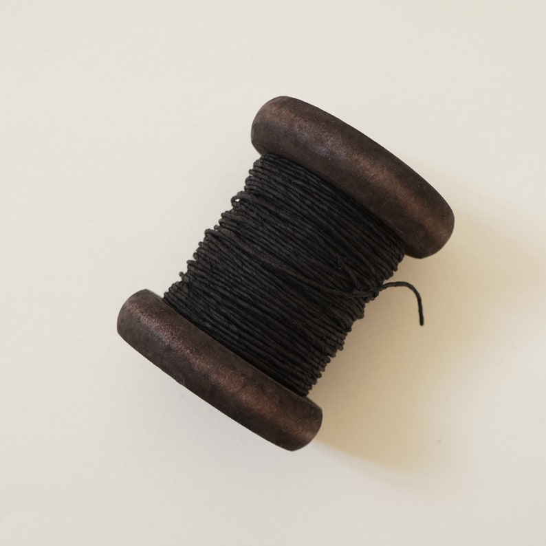 Puede incluir: Una bobina de hilo encerado negro enrollado alrededor de una bobina de madera.