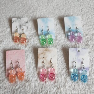 May include: Six pairs of dangle earrings featuring a turtle design. The turtles are made of translucent plastic and come in a variety of colors, including green, pink, blue, orange, and yellow.