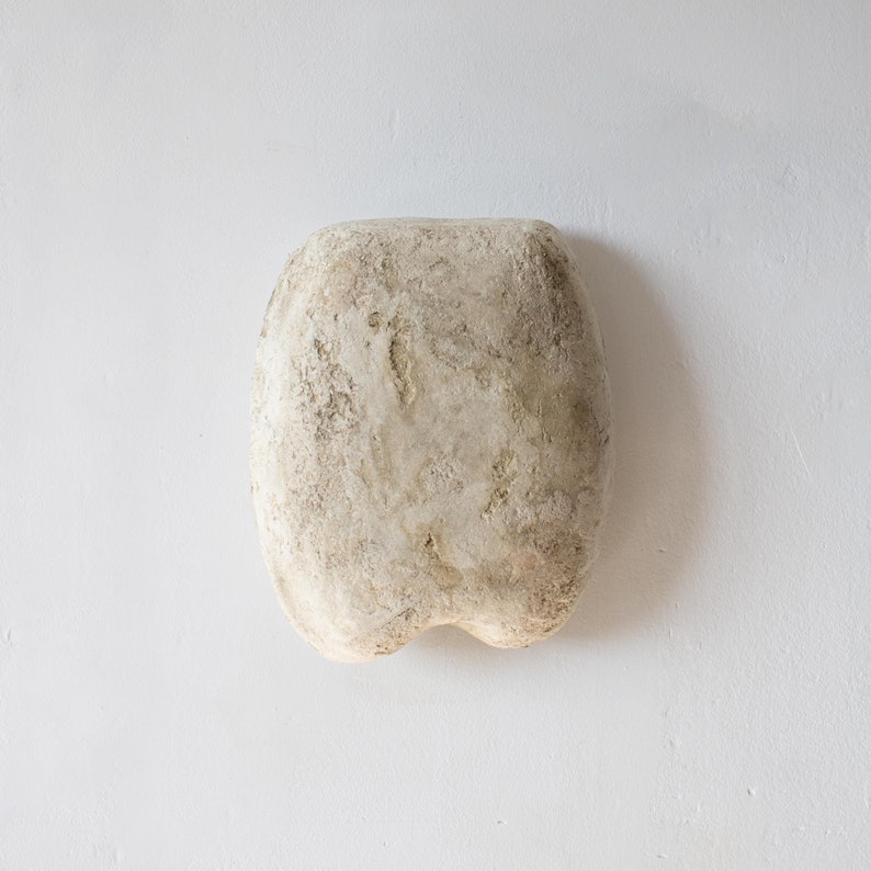 May include: An off-white, oblong sculpture with a textured surface, rounded top, and a split, rounded base. The surface has a rough, aged appearance, with variations in colour and texture. It is set against a plain white background.