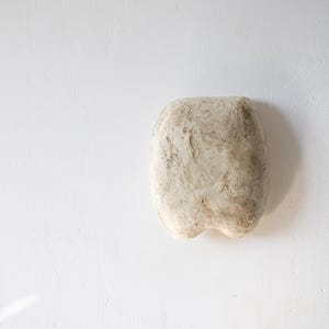 May include: A textured, off-white, rectangular sculpture with rounded edges and a slightly irregular surface. The sculpture has a rough, aged appearance, suggesting a natural or weathered material. The piece is set against a plain white background.