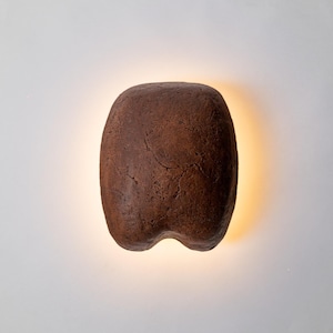 May include: A brown, organic-shaped wall sconce with a textured surface. The sconce has a rounded top and a split bottom, with a warm light emanating from behind it. The fixture is mounted on a white wall.