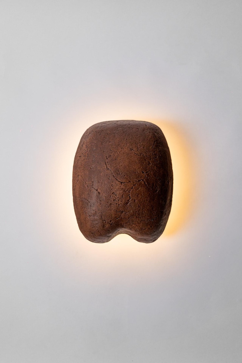 May include: A brown, organic-shaped wall sconce with a textured surface. The sconce has a rounded top and a split bottom, with a warm light emanating from behind it. The fixture is mounted on a white wall.