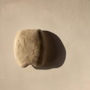 May include: A beige stone sculpture, rectangular with a rounded top and a two-lobed base. The stone has a rough, textured surface. The sculpture is set against a neutral background, with a shadow cast to the right.