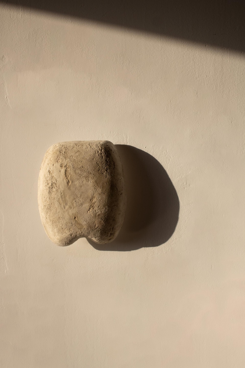 May include: A beige stone sculpture, rectangular with a rounded top and a two-lobed base. The stone has a rough, textured surface. The sculpture is set against a neutral background, with a shadow cast to the right.