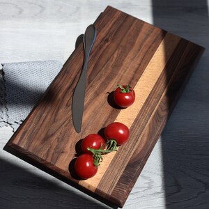 Könnte beinhalten: Ein Schneidebrett aus Holz mit einer Einlage aus hellem Holz, mit einem silbernen Messer und vier roten Kirschtomaten.