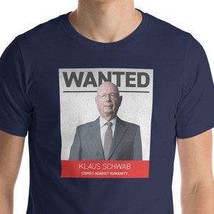 May include: A navy blue t-shirt with a white poster printed on the front. The poster says "WANTED" in red letters and features a photo of Klaus Schwab with the text "Klaus Schwab Crimes Against Humanity" underneath.