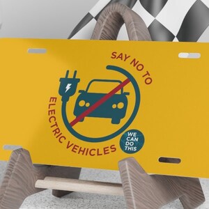 May include: A yellow license plate with a red circle and a diagonal line through it. The circle contains a black car with a blue electric plug. The text "SAY NO TO ELECTRIC VEHICLES" is written in a red and black font around the circle. The text "WE CAN DO THIS" is written in a blue font in a smaller circle below the car.