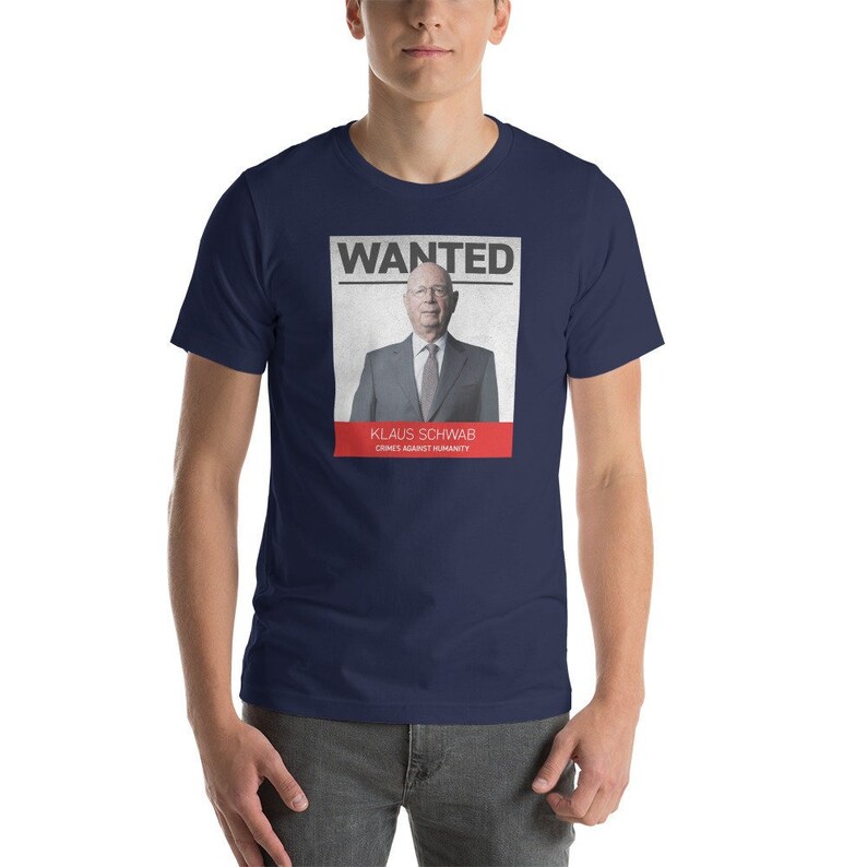 May include: A navy blue t-shirt with a white poster printed on the front. The poster says "WANTED" in red letters and features a photo of Klaus Schwab with the text "Klaus Schwab Crimes Against Humanity" underneath.