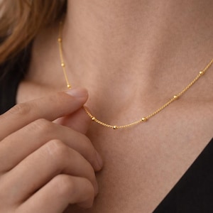 May include: Delicate gold necklace featuring small, evenly spaced beads. The necklace is displayed against a dark background, highlighting its fine details. The jewelry is being held by a hand, showcasing the necklace's length and design. A simple and elegant piece.