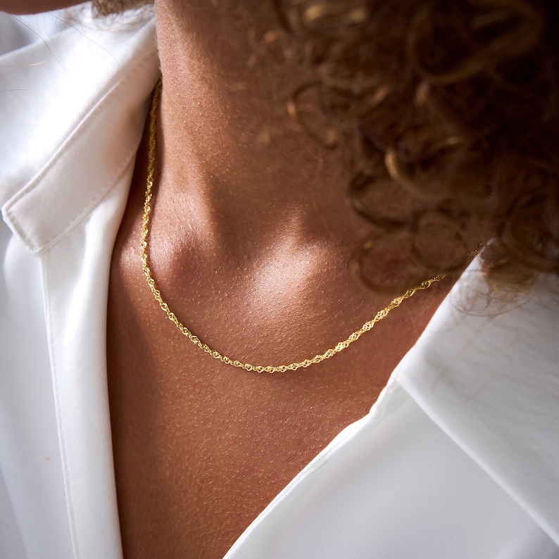 May include: A gold rope chain necklace on a person's neck. The chain is thin and delicate, and it is worn against a white shirt.