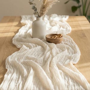 May include: A cream-coloured, textured table runner draped across a wooden table. A white vase holds dried flowers and greenery. A small wooden bowl contains dried orange slices, creating a neutral, natural aesthetic.