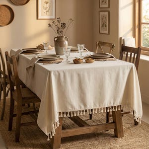 May include: A dining room with a wooden table set for a meal. The table is covered with a cream tablecloth with tassel trim. Brown plates, glasses, and a vase with dried flowers are on the table. Wooden chairs surround the table.