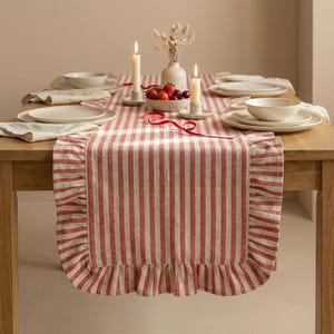 May include: A dining table set for a meal, featuring a red and white striped table runner with a ruffled edge. The table is set with white plates, bowls, and napkins. Lit candles and a bowl of fruit add to the ambiance.