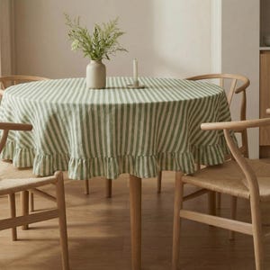 May include: A round dining table set with a green and white striped tablecloth featuring a ruffled edge. The table is surrounded by four wooden chairs with woven seats. A vase with greenery and a candle are on the table.