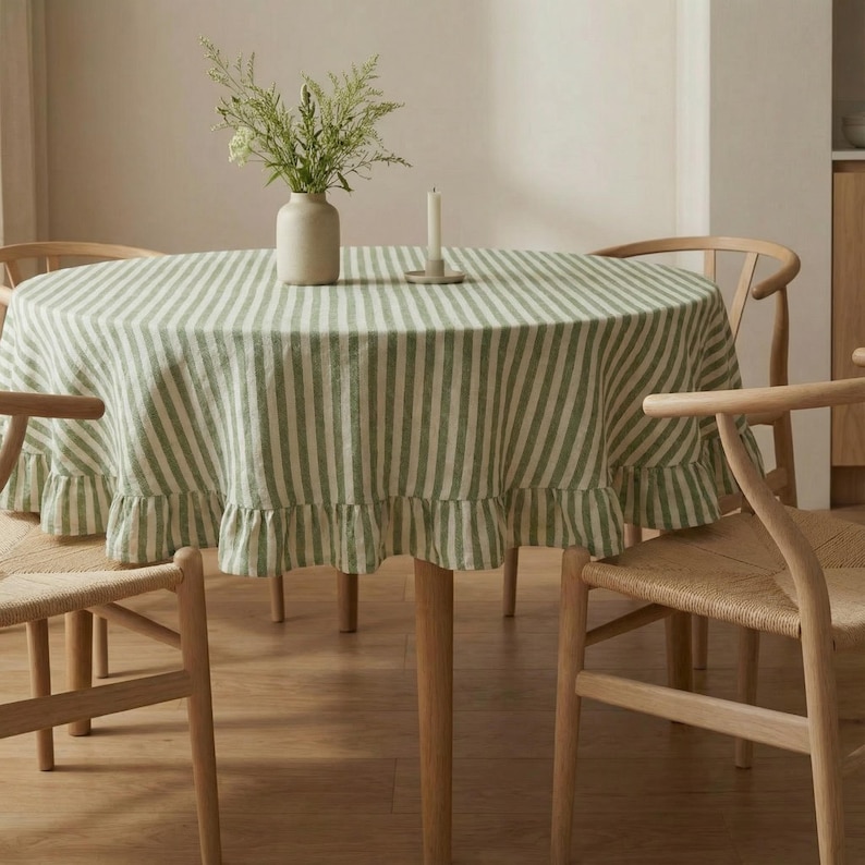 May include: A round dining table set with a green and white striped tablecloth featuring a ruffled edge. The table is surrounded by four wooden chairs with woven seats. A vase with greenery and a candle are on the table.