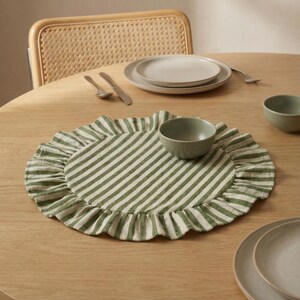 May include: A round, green and white striped placemat with a ruffled edge. A small, green bowl sits on the placemat, with a light-colored plate and bowl in the background. The placemat is on a wooden table.