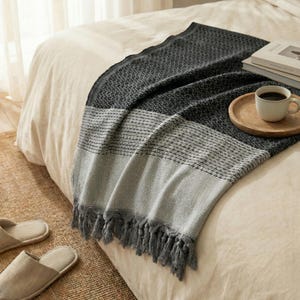 May include: A gray and white woven throw blanket with a geometric pattern and fringe detail. The blanket is draped over a bed with a neutral-colored comforter. A wooden tray with a coffee cup and books are on the bed.