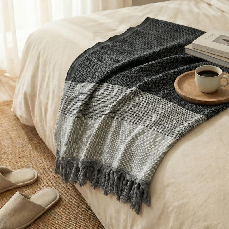 May include: A gray and white woven throw blanket with a geometric pattern and fringe detail. The blanket is draped over a bed with a neutral-colored comforter. A wooden tray with a coffee cup and books are on the bed.