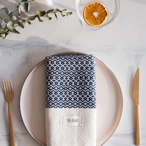 May include: A folded navy and white patterned towel with fringe, placed on a white plate. A gold fork and knife are on either side. A glass bowl with a dried orange slice is in the background.