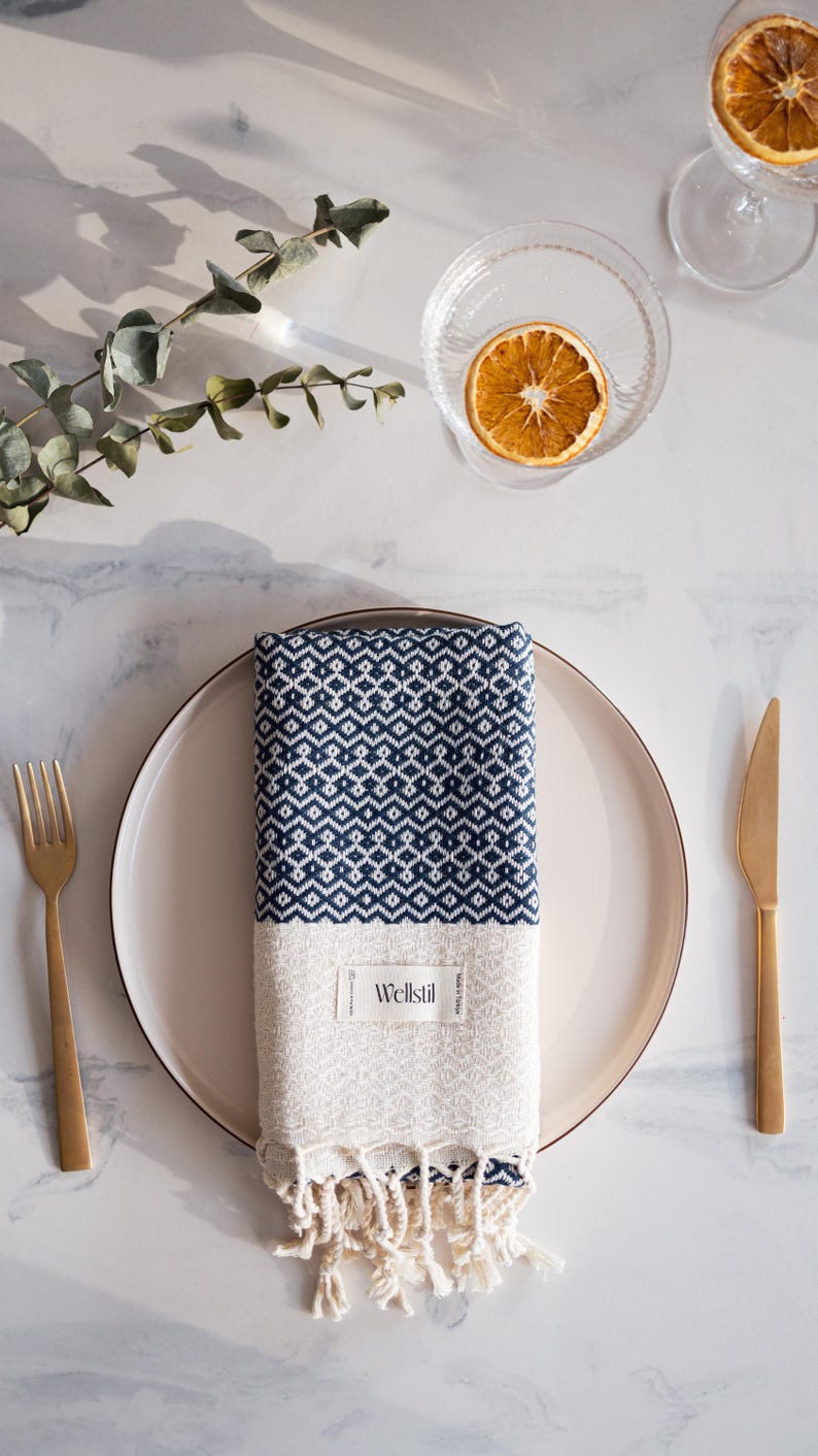 May include: A folded navy and white patterned towel with fringe, placed on a white plate. A gold fork and knife are on either side. A glass bowl with a dried orange slice is in the background.
