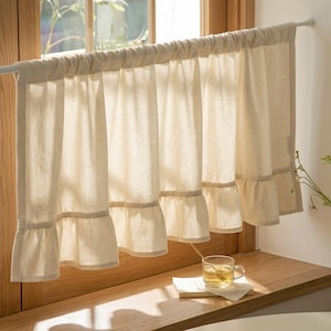 Peut inclure: Rideau de fenêtre crème avec un bord inférieur à volants. Le rideau est fait d'un tissu léger et est suspendu à une tringle blanche. Le rideau est devant une fenêtre avec de la lumière naturelle.