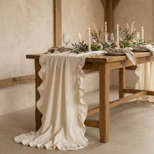 May include: A rustic wooden table is set for a formal dinner. A cream-colored ruffled table runner drapes over the table. The table is decorated with candles, greenery, wine glasses, and plates, creating an elegant and inviting atmosphere.