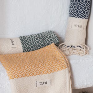 May include: Three rolled and folded patterned towels in shades of yellow, green, and navy blue. The towels have a geometric design and fringe. Each towel has a white label that reads "Wellstil". A small potted cactus is in the background.