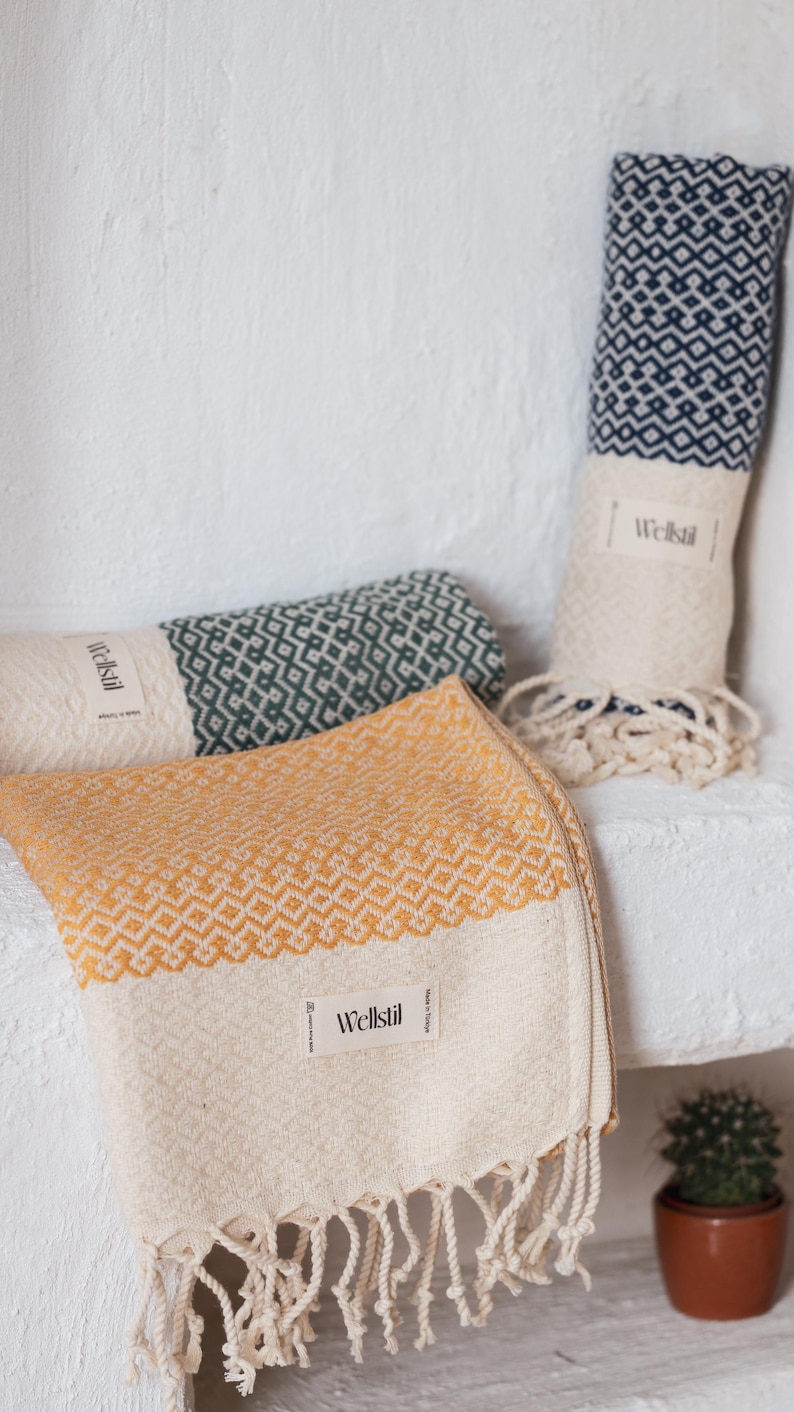 May include: Three rolled and folded patterned towels in shades of yellow, green, and navy blue. The towels have a geometric design and fringe. Each towel has a white label that reads "Wellstil". A small potted cactus is in the background.
