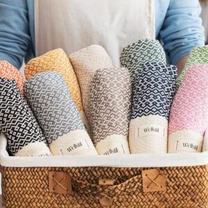 May include: A wicker basket containing rolled towels in a variety of colours, such as black, grey, yellow, beige, brown, navy, and pink. Each towel features a geometric design and a label reading "Wellstil".