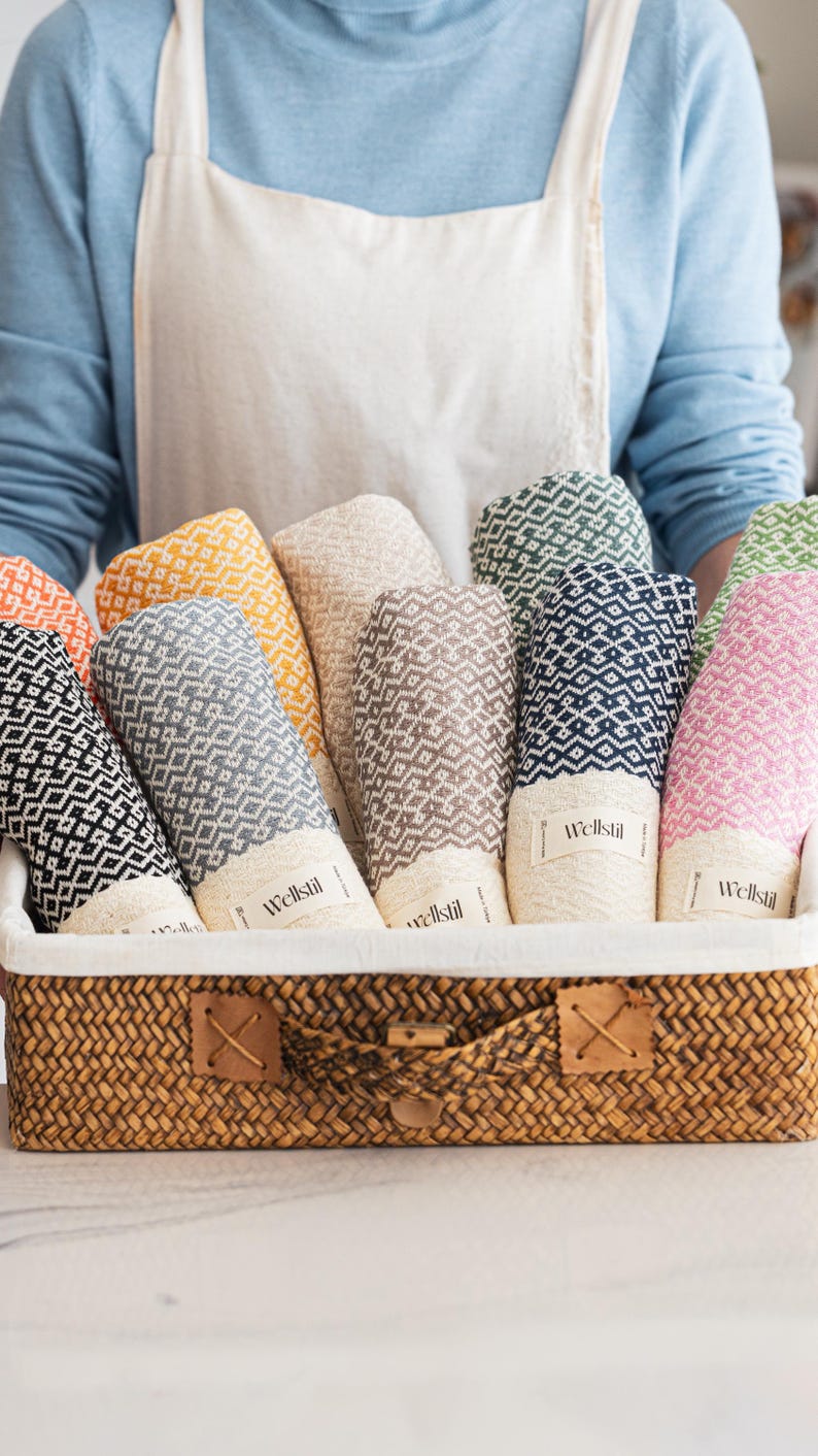 May include: A woven basket filled with rolled-up towels in various colors, including black, gray, yellow, beige, brown, navy, and pink. Each towel has a geometric pattern and a label that reads "Wellstil".