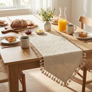 May include: A wooden dining table set for breakfast, featuring a woven cream-colored table runner with tassels. The table is set with plates of pastries, bread, butter, jam, coffee, and orange juice. A vase of flowers adds a decorative touch.