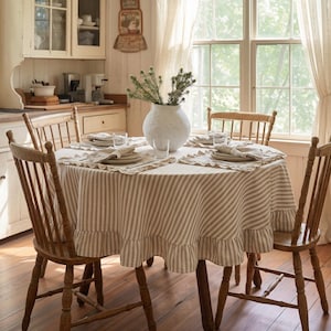 Op de afbeelding: Een ronde eettafel voor vier personen met een gestreept tafelkleed en een geschulpte rand. De tafel is omringd door houten stoelen. Een witte vaas met groen staat in het midden. De kamer heeft een rustieke, boerderijstijl.