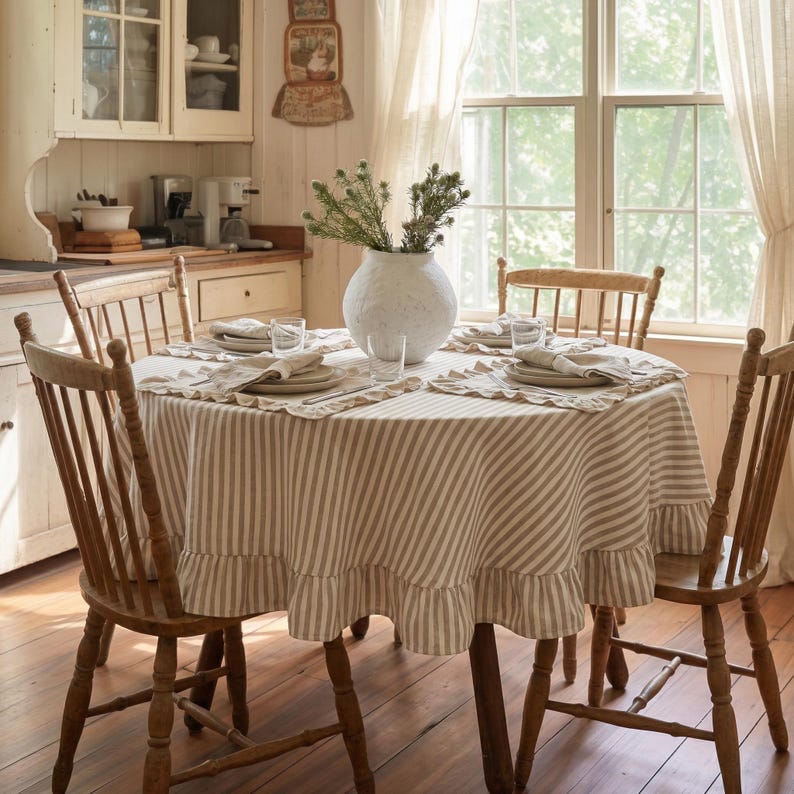 Op de afbeelding: Een ronde eettafel voor vier personen met een gestreept tafelkleed en een geschulpte rand. De tafel is omringd door houten stoelen. Een witte vaas met groen staat in het midden. De kamer heeft een rustieke, boerderijstijl.