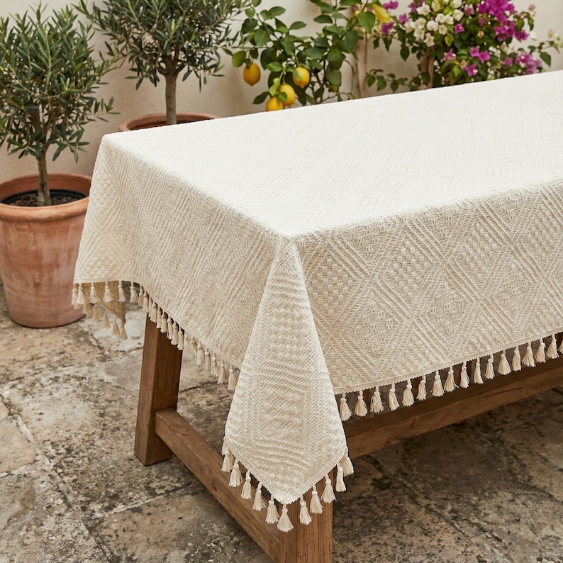 K&ouml;nnte beinhalten: Cremefarbene rechteckige Tischdecke mit geometrischem Rautenmuster und Fransenquasten. Die Tischdecke ist &uuml;ber einem Holztisch drapiert und erzeugt einen strukturierten Look. Ideal f&uuml;r Ess- oder Au&szlig;enbereiche.