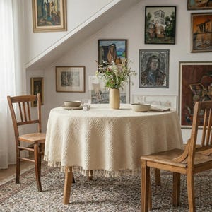 Op de afbeelding: Een ronde, crèmekleurige tafelkleed met een textuurpatroon en kwastjesrand bedekt een houten tafel. Twee houten stoelen staan rond de tafel. Een vaas met bloemen staat in het midden, met kunstwerken aan de muur op de achtergrond.