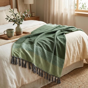 May include: A green woven throw blanket with a textured design and gray tassels. The blanket is draped over a bed with a cream-colored comforter. The throw has a striped pattern in varying shades of green.