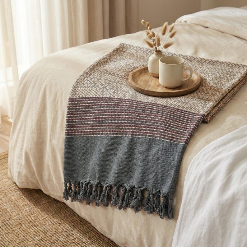 May include: A woven throw blanket with a geometric pattern in beige, red, and gray, draped over a bed. A wooden tray holds a white mug and small vase with dried flowers. The blanket has a fringed edge.