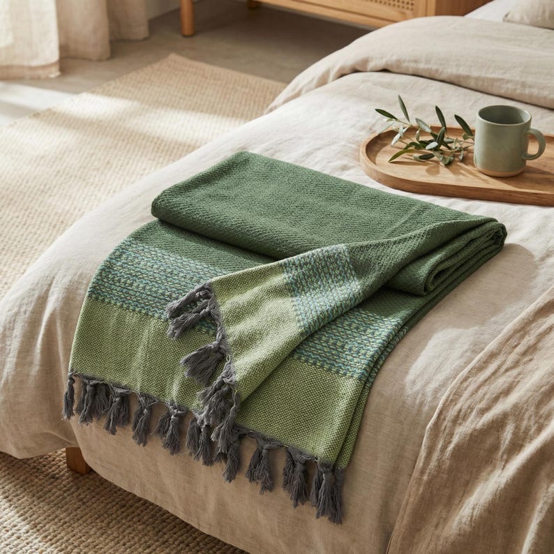 May include: A green woven throw blanket with a textured pattern and gray tassels. The blanket is folded on a bed with a neutral-colored duvet. A wooden tray with a mug and greenery sits on the bed.