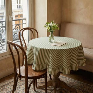 May include: A round wooden table with a green and white striped tablecloth and a ruffled edge. Two wooden chairs are positioned near the table. A vase of flowers sits on the table. The room has a window and a beige sofa.