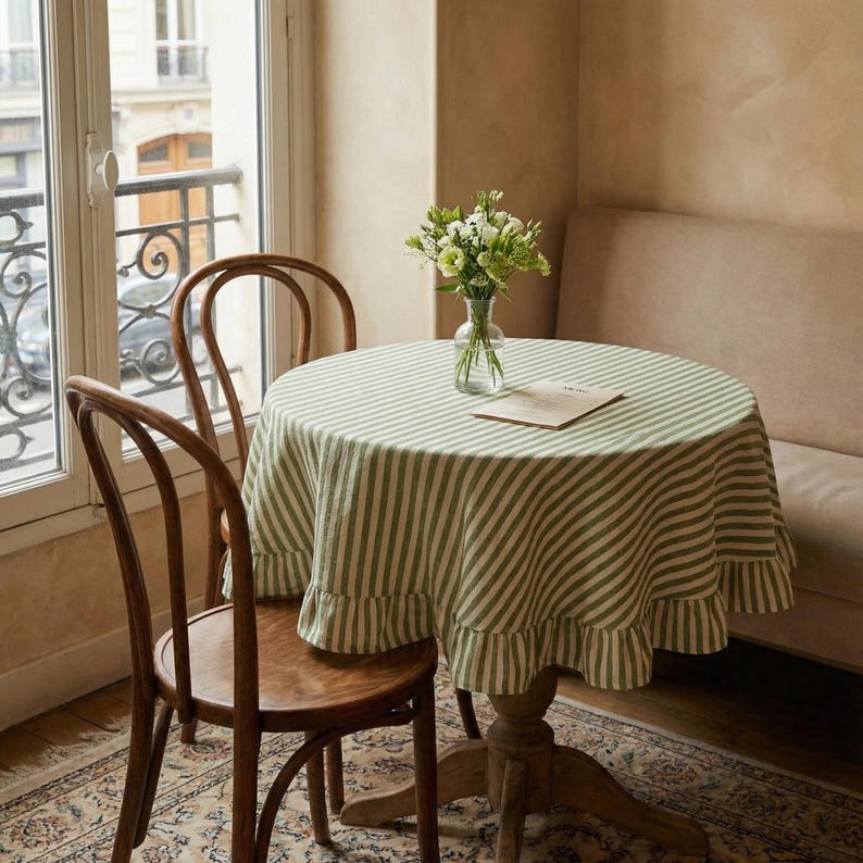 May include: A round wooden table with a green and white striped tablecloth and a ruffled edge. Two wooden chairs are positioned near the table. A vase of flowers sits on the table. The room has a window and a beige sofa.