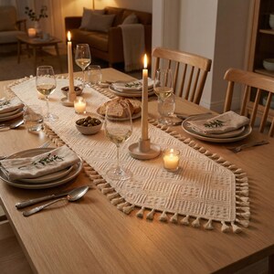 May include: A dining table set for a meal, featuring a cream-colored table runner with a geometric pattern and tassels. The table is set with plates, glasses, candles, and silverware, creating a warm and inviting atmosphere.