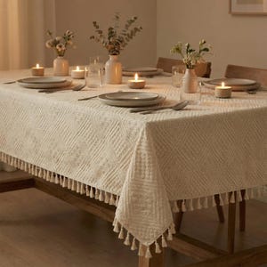K&ouml;nnte beinhalten: Ein cremefarbenes Tischtuch mit einem strukturierten geometrischen Muster und Fransenbesatz bedeckt einen Holztisch. Der Tisch ist mit Tellern, Besteck, Kerzen und Vasen mit Blumen gedeckt, was eine warme Atmosph&auml;re schafft.