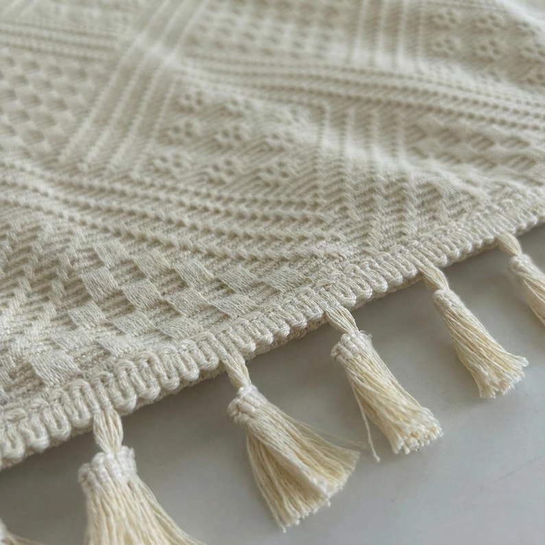 K&ouml;nnte beinhalten: Cremefarbene gewebte Decke mit geometrischem Muster und Fransenquasten am Rand. Die Decke hat ein strukturiertes Design mit Rauten- und Fischgr&auml;tmustern, geeignet f&uuml;r die Inneneinrichtung.