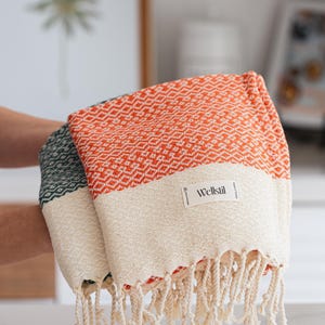 May include: Two folded Turkish towels. One is orange with a white band and diamond pattern, the other is green with a white band. Both have fringe and a "Wellstil" label.