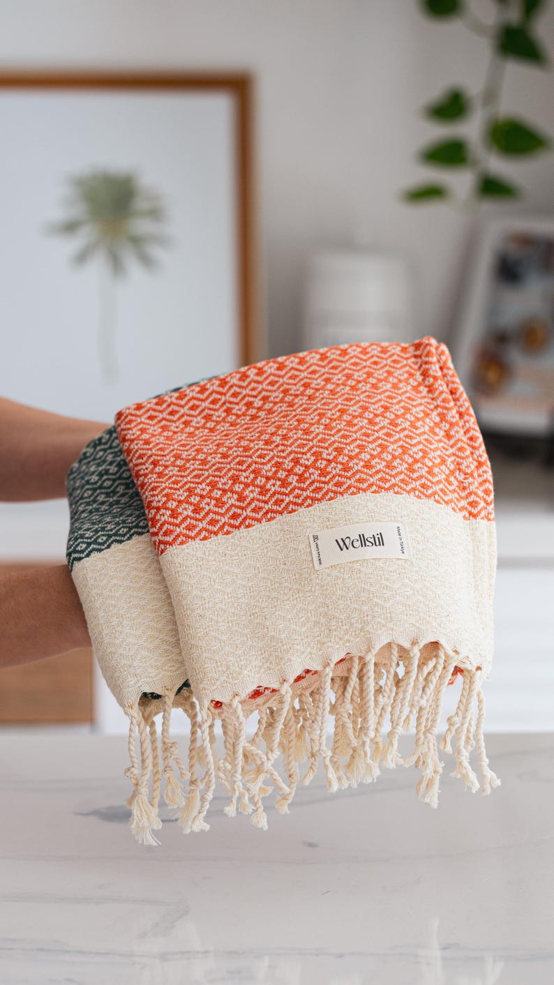 May include: Two folded Turkish towels. One is orange with a white band and diamond pattern, the other is green with a white band. Both have fringe and a "Wellstil" label.