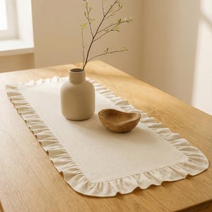 May include: A rectangular off-white table runner with a ruffled edge sits on a wooden table. A small, beige vase holds a sprig of greenery. A small, wooden bowl is also on the runner.